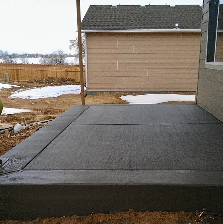 New concrete driveway pad in front of tan garage with wooden fence and patches of snow in background