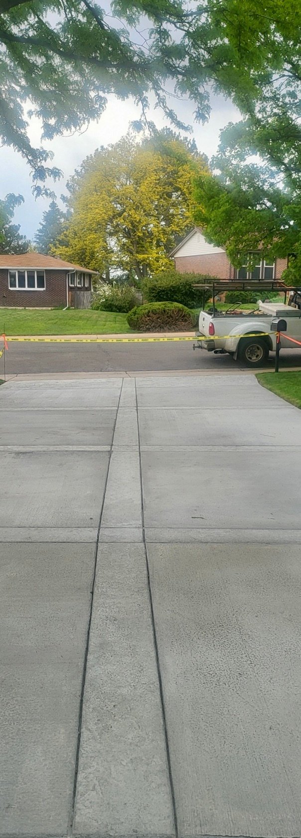 Residential driveway with yellow caution tape, brick house, work trailer, and mature trees in background