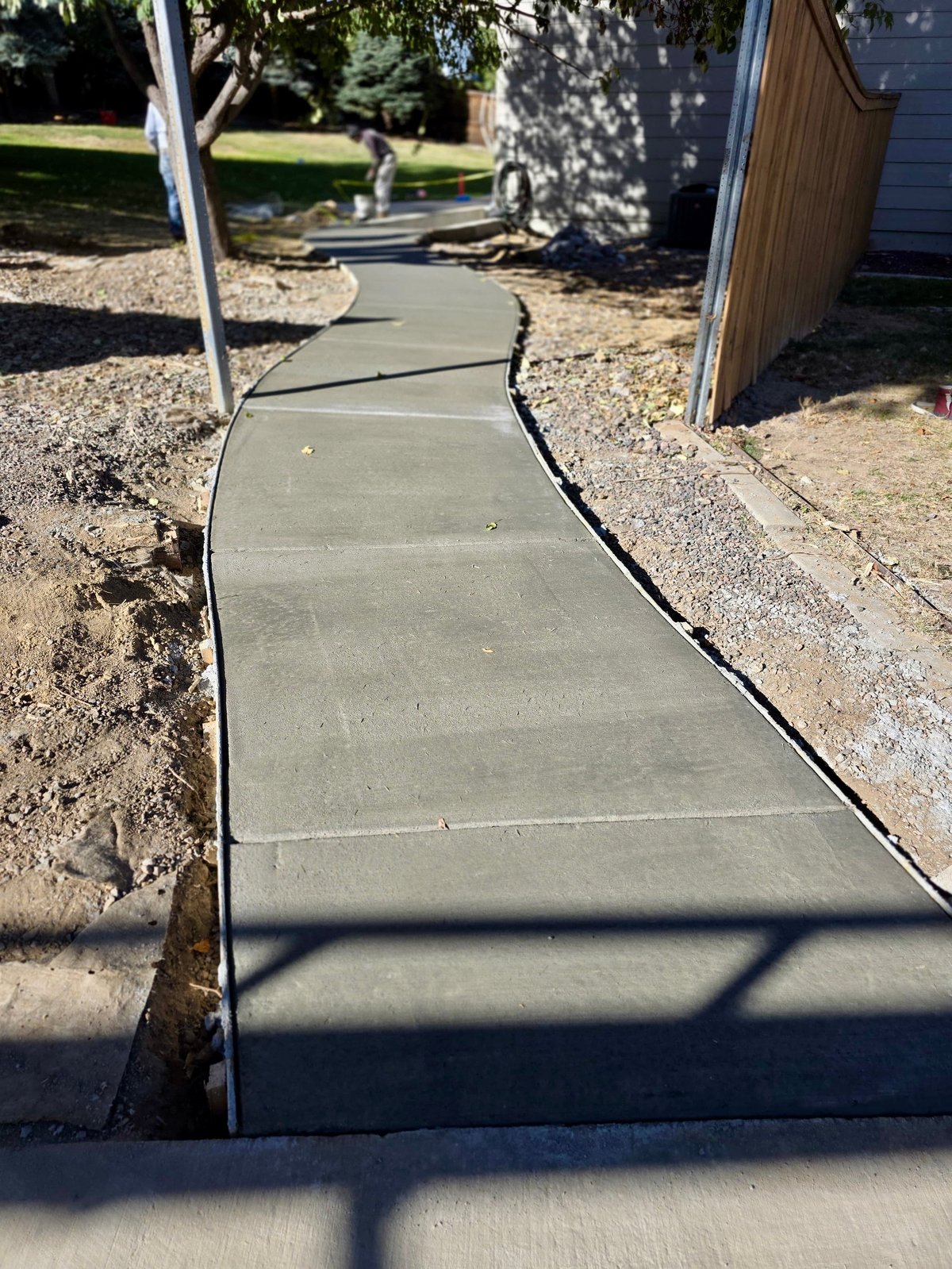 Newly poured concrete pathway winding through a backyard with landscaping elements and wooden structures visible on sides