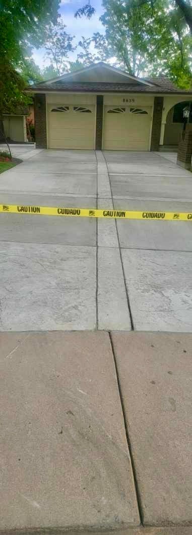 Driveway with yellow caution tape across concrete slabs in front of brick garage with two doors and large trees overhead