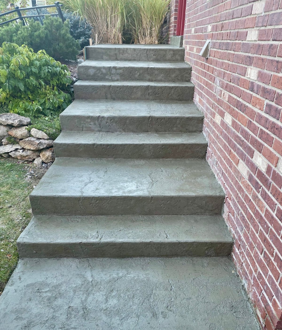 Outdoor concrete steps leading upward beside a red brick wall, with ornamental grasses and plants visible in the background