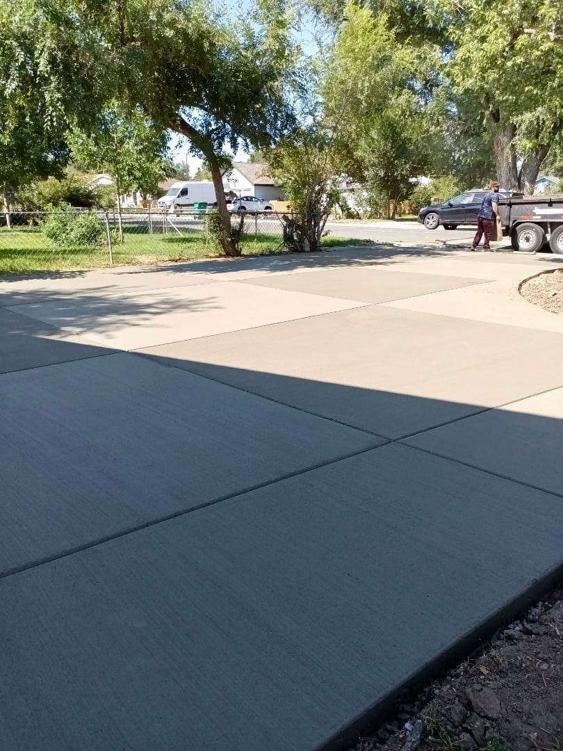 Newly poured concrete driveway in residential neighborhood with trees, parked vehicles, and houses visible in background