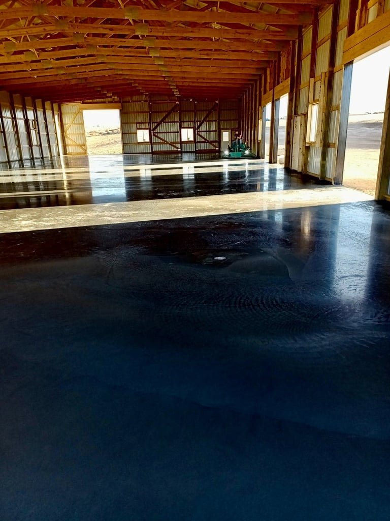 Large empty barn interior with polished dark concrete floor, wooden beam ceiling, and open doors showing rural landscape